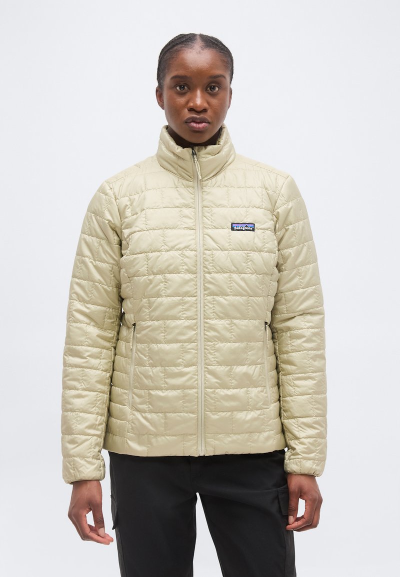 Άτομο που φοράει ανοιχτό μπεζ μπουφάν με καπιτονέ ύφασμα και ψηλό γιακά, καθώς και μαύρο παντελόνι, στέκεται μπροστά σε απλό λευκό φόντο.