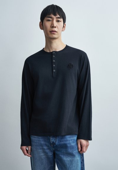 Μαύρο μακρυμάνικο πουκάμισο henley με στρογγυλεμένο λαιμόκοψη, πέντε κουμπιά και κεντημένο λογότυπο στο αριστερό στήθος, συνδυασμένο με μπλε τζιν.