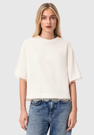 Kremlfarvet kortærmet top med blondedetaljer ved kant og ærmer, med en afslappet pasform og blød tekstur. Kombination med lyseblå jeans.