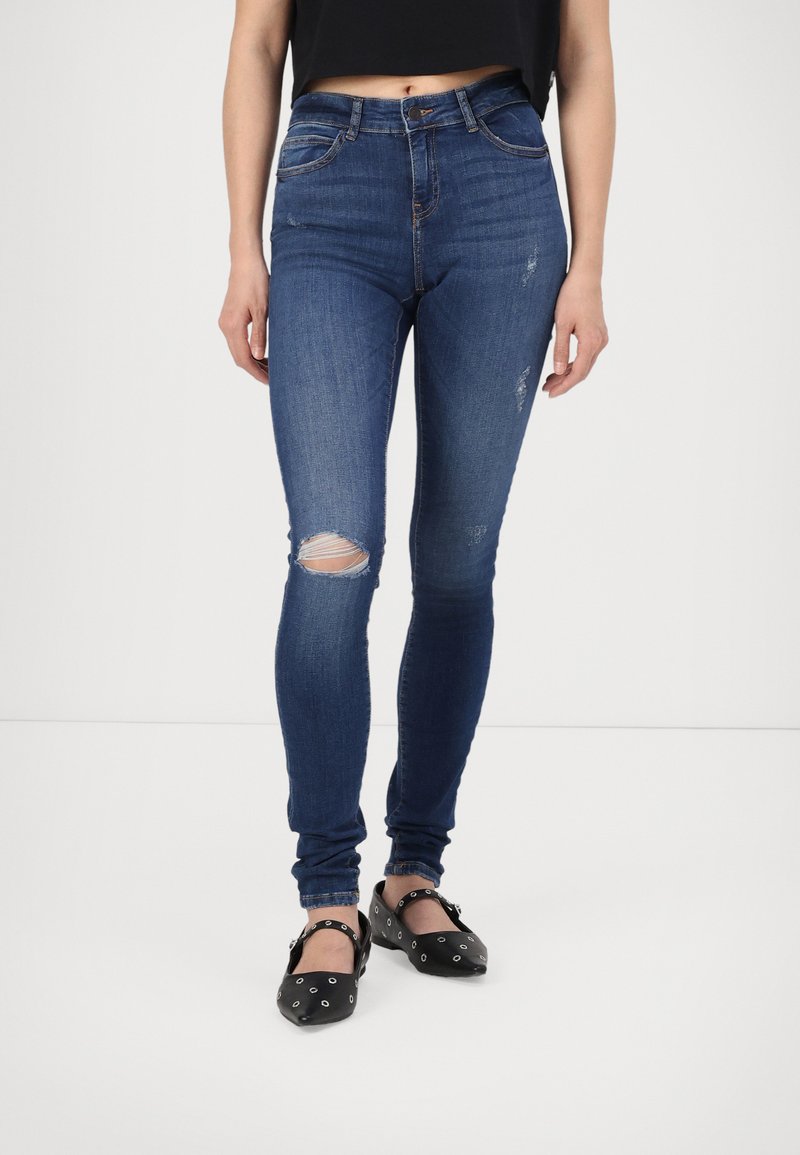 Högmidjade blå jeans med smal passform, som har slitna knäskärningar och en mjuk denimstruktur. Bärs med svarta skor.