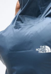 Άτομο που φοράει μπλε μπουφάν με κουκούλα με το λογότυπο The North Face να φαίνεται στην πίσω περιοχή του ώμου.