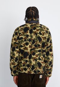 Veste en polaire avec un motif léopard vert, marron et noir. Col haut et surface texturée avec une coupe décontractée.