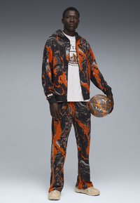 Atlet i en sort og orange marmorering træningsdragt med matchende bukser, hvid grafisk t-shirt og beige sneakers, der holder en basketball.