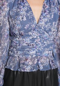 Bluse med blomsterprint i lyseblå, lilla og hvid. Den har dyb V-hals, knaplukning foran og flæset talje med gennemsigtige lange ærmer.