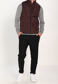 Ciemno-bordowa wypełniona kamizelka z wysokim kołnierzem, zapinana na zamek, w połączeniu z szarym, długim rękawem, czarnymi spodniami i białymi sneakersami.