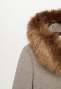 Abrigo de lana beige que cuenta con un gran y suave cuello de piel sintética marrón. Los bordes del cuello son voluminosos y texturizados, contrastando con la tela lisa del abrigo.