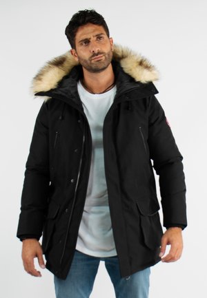 Parka noire avec une capuche en fourrure, fermetures éclair et boutons-pression, grandes poches avant et tissu résistant à l'eau. Coupe classique avec manches réglables.