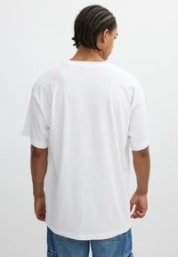 Hombre con trenzas en cornrows que lleva una camiseta blanca de gran tamaño y shorts de cargo de mezclilla azules, de espaldas contra un fondo neutro.