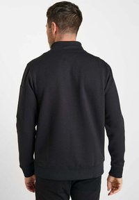Sweatshirt noir en coton, avec un col montant et des poignets côtelés. Le dos a une finition texturée et une poche latérale.