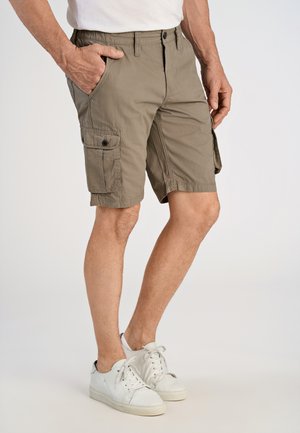 Mężczyzna w khakowych spodenkach cargo i białych tenisówkach, stojący z jedną ręką w kieszeni na jasnym tle.