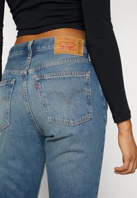 Personne portant un jean Levi's bleu clair et un top court noir à manches longues, vu de dos de la taille jusqu'à mi-cuisse.