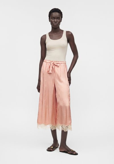 Γυναίκα με κοντά μαλλιά που φοράει μπεζ αμάνικη μπλούζα, ροζ φαρδιά cropped παντελόνια με δαντελένιο τελείωμα και μαύρα σανδάλια με χρυσή λεπτομέρεια.