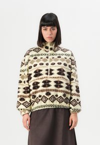 Vrouw met steil donker haar die een patroonrijke crèmekleurige fleece pullover met geometrische ontwerpen en een donkere rok draagt, tegen een effen achtergrond.