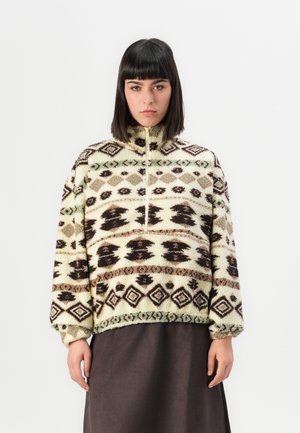 Woman with straight dark hair wearing a patterned cream fleece pullover with geometric designs and a dark skirt against a plain background.