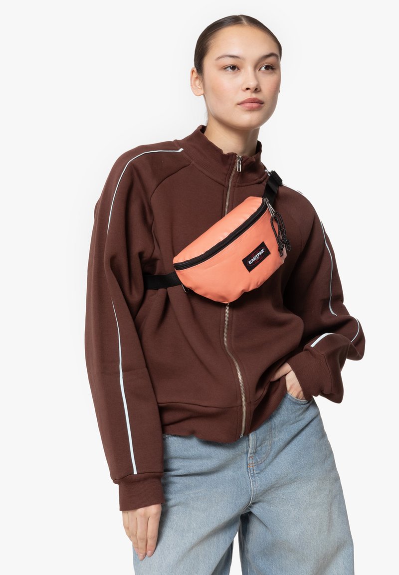 Korallfärgad midjeväska med dragkedjestängning och svart rem, bärs över en brun zip-up tröja med vita accentlinjer, i kombination med ljusblå jeans.