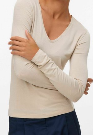 Personne portant un haut beige à manches longues côtelé avec un col en V, tenant le bras opposé croisé sur la poitrine sur fond blanc.