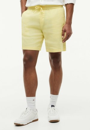 Mann trägt blassgelbe Shorts mit Kordelzug, weiße Sportsocken und weiße Sneakers, steht vor einem einfarbigen Hintergrund.