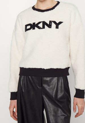 Persoon die een witte, pluizige DKNY-trui draagt met zwarte manchetten en zwarte nepleren broek, zichtbaar van nek tot halverwege de dij.