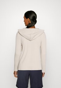 Beige gesteppter Langarm-Top mit Kapuze; verfügt über einen strukturierten Stoff und eine lässige Passform, kombiniert mit navy Shorts.