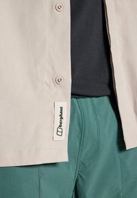 Beige button-up shirt with a smooth texture, featuring a tag with "berghaus" logo. Worn over a dark shirt and paired with green shorts.