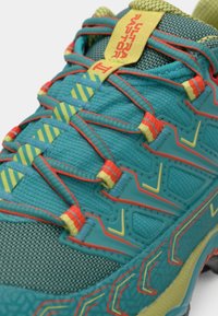 Sapatilha de corrida em turquesa com parte superior em malha texturizada, atacadores amarelos e vermelhos, marcação em destaque e sola acolchoada com um padrão geométrico.