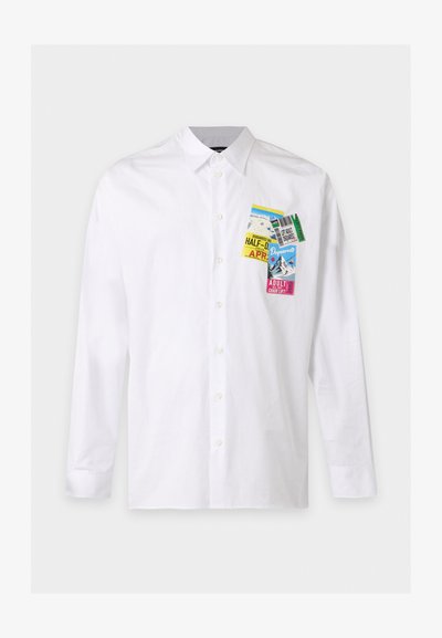 Chemise blanche à manches longues boutonnée avec des écussons graphiques colorés de tickets de remontée mécanique sur la partie gauche de la poitrine.