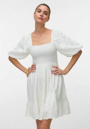 Kobieta w białej sukience z bufkami na rękawach, z kwadratowym dekoltem i marszczonym górnym stanem, pozująca z rękami na biodrach na białym tle.