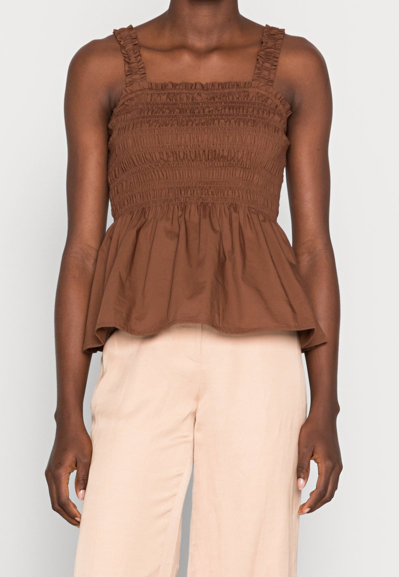 Haut péplum smocké marron avec un décolleté carré et des bretelles volantées, fabriqué en tissu léger. Pantalon beige clair complétant la tenue.