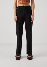 Black, high-waisted trousers with an elastic waistband, featuring vertical pleats and smooth texture. Paired with chunky sandals.