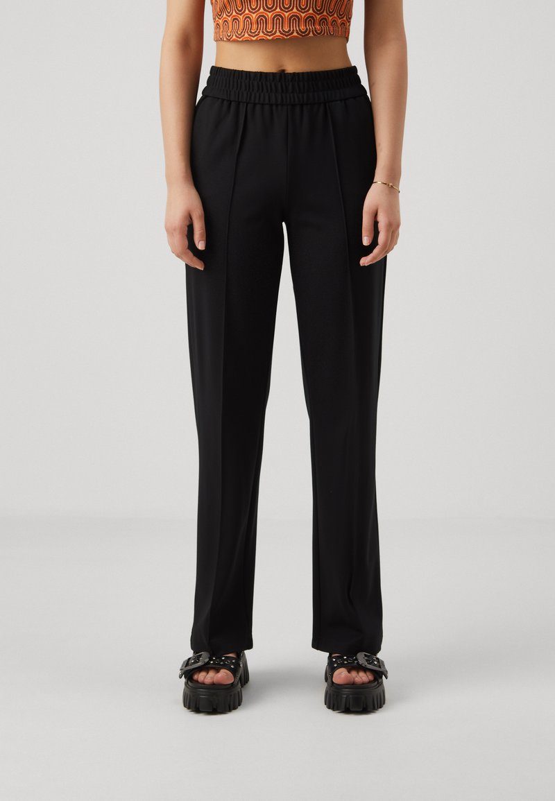 Black, high-waisted trousers with an elastic waistband, featuring vertical pleats and smooth texture. Paired with chunky sandals.