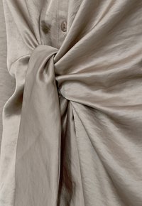 Beige, zijden stof met een knoopdetail, met een glad, textuur oppervlak en subtiele glans, wat een gedrapeerd uiterlijk creëert. Knopen zichtbaar.