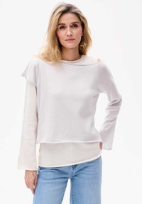 Langärmliges, leichtes weißes Oberteil mit weitem Rundhalsausschnitt, gespaltenem Schulterdesign und einem geschichteten Look, kombiniert mit hellblauen Jeans.