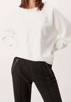 Sudadera blanca con cuello redondo y mangas raglán, combinada con pantalones negros que tienen cremalleras en los bolsillos laterales.