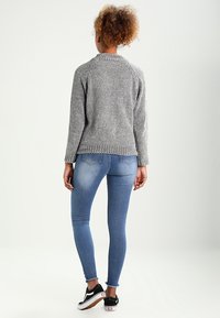 Grau gestrickter Pullover mit ribbeltem Saum, kombiniert mit blauen Skinny-Jeans und schwarzen Sneakers, von hinten betrachtet.