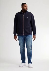 Námořnicky modrá mikina na zip s texturovanou látkou, přední kapsou a kontrastním zipem. Kombinována s světle modrými džínami a bílými teniskami.