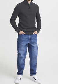 Tmavě šedý texturovaný svetr s límcem na zip, v kombinaci se světle modrými džíny. Obuv má bílou barvu s černými akcenty.