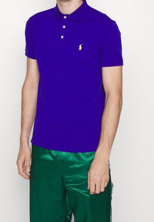 Homme portant un polo bleu royal avec un logo jaune et un pantalon vert brillant, debout devant un fond blanc uni.