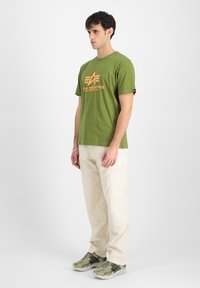 Grøn bomulds t-shirt med orange logo, parret med beige cargo bukser og grå-grønne sneakers med textured overflade og mørke accenter.
