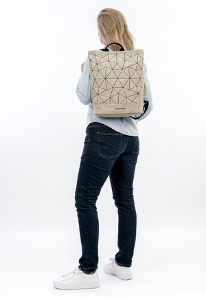 Frau mit langen blonden Haaren, die ein hellblaues Hemd, dunkle Jeans und weiße Turnschuhe trägt, und einen beigen Rucksack mit geometrischem Muster von SURI FREY bei sich hat.