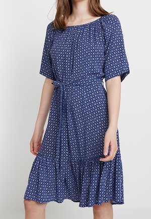 Kobieta w niebieskiej sukience do kolan z białym kwiatowym wzorem, krótkimi rękawami, taliowanym paskiem i falbaną na dole.