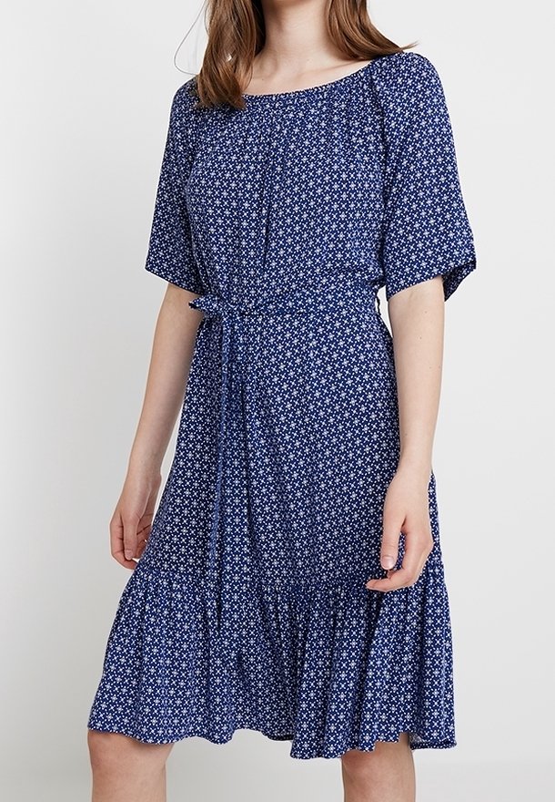Femme portant une robe bleue longueur genou avec un motif floral blanc, manches courtes, ceinture nouée à la taille et ourlet à volants.