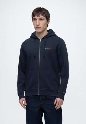 Ung mann iført en marineblå hettegenser med glidelås og frontlommer med "Tommy Jeans New York"-logo på brystet, kombinert med mørkeblå jeans.