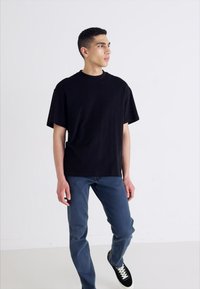 Jonge man staande, gekleed in een zwart t-shirt, blauwe jeans en zwarte sneakers met witte veters, die naar links kijkt tegen een eenvoudige witte achtergrond.