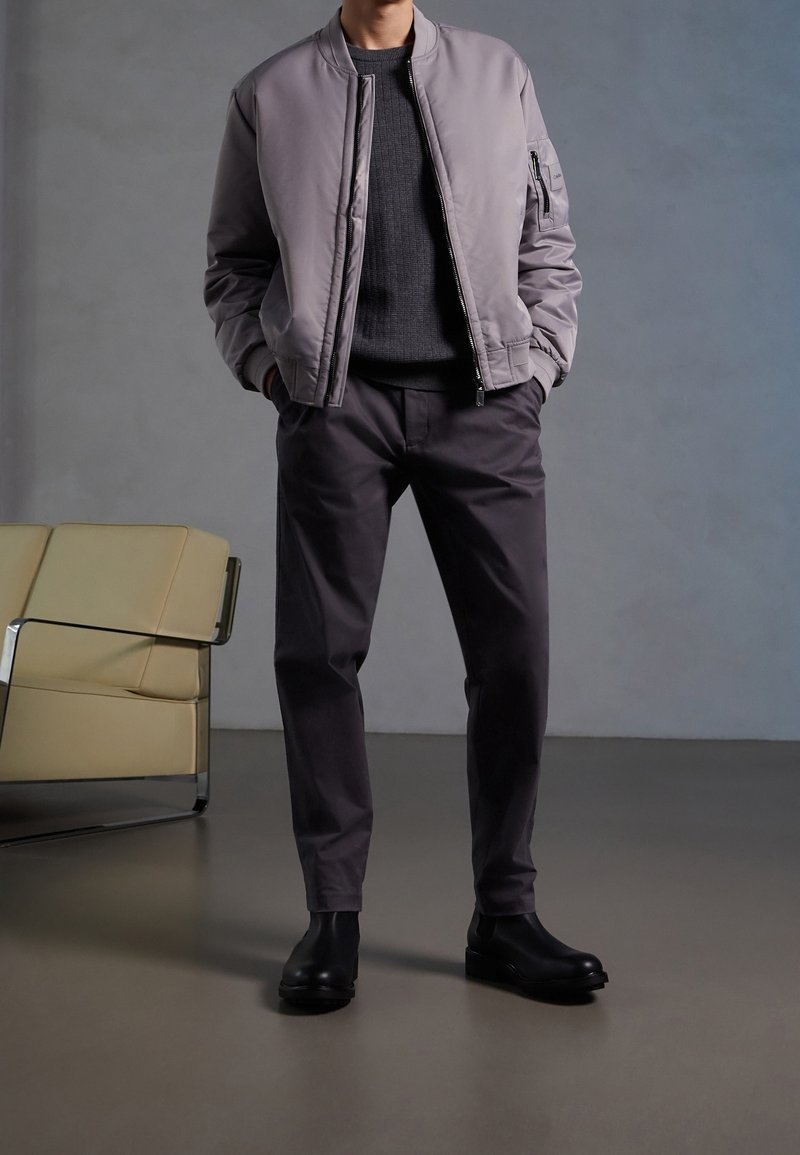 Giacca bomber grigio chiaro sopra un maglione scuro e strutturato, abbinata a pantaloni grigio scuro e stivali neri alla caviglia. Tessuto morbido e design semplice.