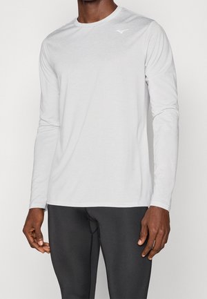 Mann trägt einfaches weißes langärmliges Sportshirt und schwarze Leggings, steht mit entspannten Armen an den Seiten vor einfarbigem Hintergrund.