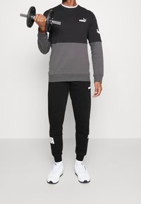 Černá a šedá sportovní mikina s dlouhými rukávy, doplněná černými tepláky s bílým logem. Drží černou činkou.