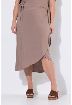 Kvinde iført en taupe indpakningsnederdel kombineret med brune åbne platformhæle, stående mod en hvid væg.