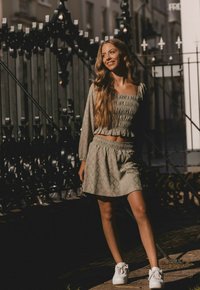 Licht olijfgroen twee-delig outfit met een gesmockte crop top met lange mouwen en een bijpassende flared rok, gecombineerd met witte sneakers.