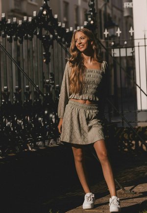 Licht olijfgroen twee-delig outfit met een gesmockte crop top met lange mouwen en een bijpassende flared rok, gecombineerd met witte sneakers.