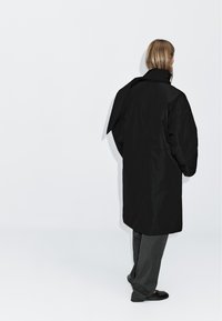 Black oversized coat with a high collar and long sleeves; features a back tie detail. Worn over gray trousers and black shoes.